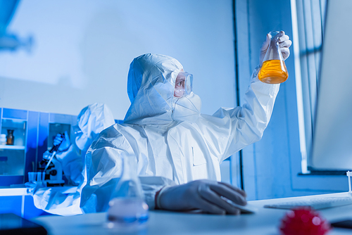 bioengineer in hazmat suit looking at flask with orange liquid in flask