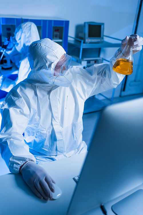 scientist in hazmat suit holding flask with orange liquid near blurred monitor in lab