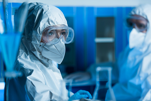 woman in goggles, medical mask and hazmat suit near scientist on blurred background