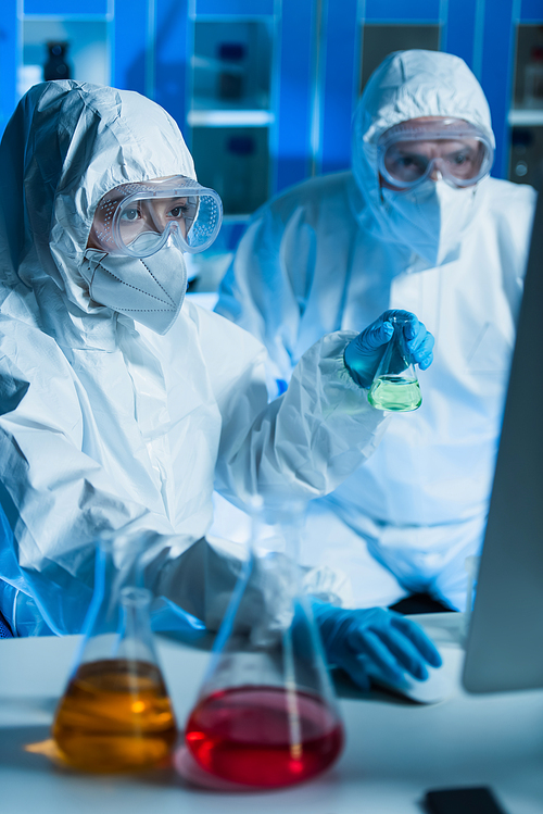 scientists in hazmat suits working in laboratory near blurred flasks with colorful liquid