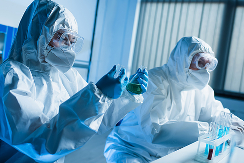 scientist in hazmat suit holding medical flasks while working with colleague in lab