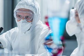 man in goggles and hazmat suit working near blurred colleague in laboratory