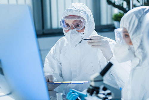 virologist in hazmat suit holding clipboard and pointing at blurred monitor near colleague
