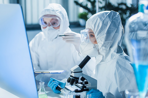 woman in hazmat suit working with microscope near computer monitor and blurred colleague
