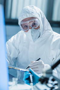 virologist in hazmat suit and goggles looking at camera on blurred foreground