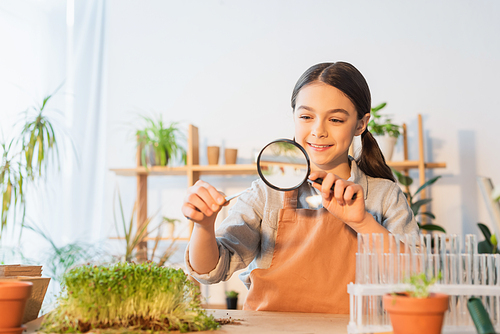 Smiling girl holding plant in tweezers and magnifying glass at home
