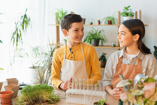 Preteen kids in aprons looking at each other near test tubes and plants at home