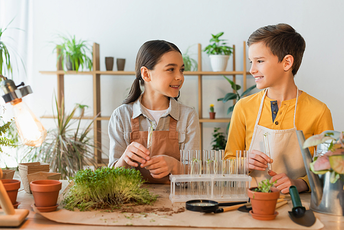 Smiling girl in apron looking at friend near glass test tubes and plants at home