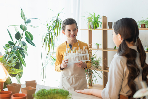 Smiling boy holding test tubes near blurred friend and plants at home