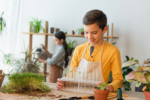 Smiling boy holding test tubes with plants near gardening tools on table at home