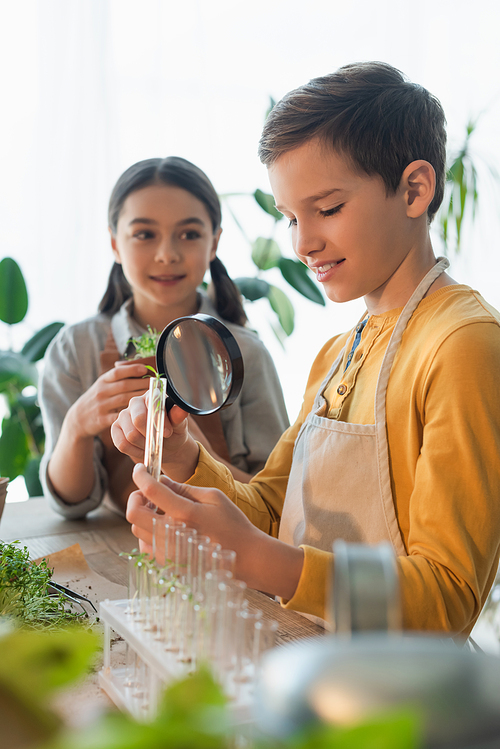 Cheerful boy holding magnifying glass and test tube near blurred friend and plants at home