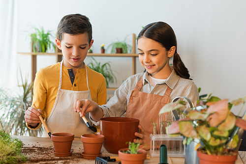 Smiling girl pouring ground in flowerpot near friend and plants at home
