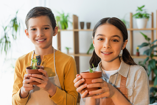 Smiling kids in aprons holding flowerpots with plants at home