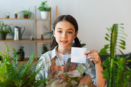 Preteen girl in apron holding sticky note near blurred plant at home