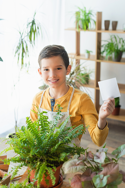 Smiling kid holding sticky note near green plants at home