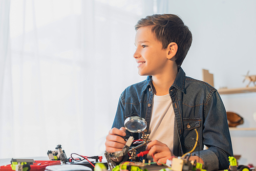 Smiling preteen boy holding magnifying glass near robotics model at home