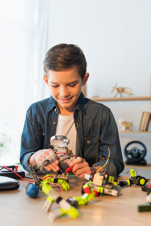 Cheerful boy holding magnifying glass near robotics model at home