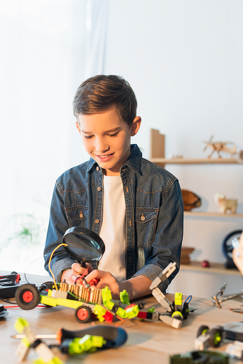 Smiling boy holding magnifying glass near blurred robotic model at home