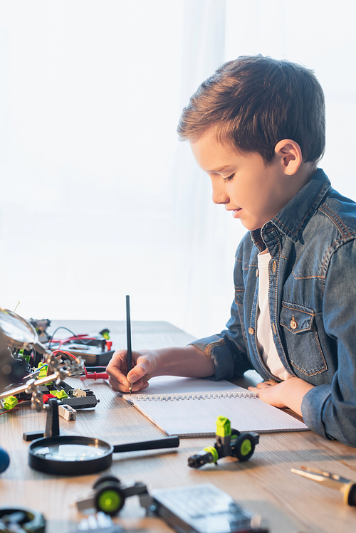 Side view of preteen boy writing on notebook near magnifying glass and robotic model at home