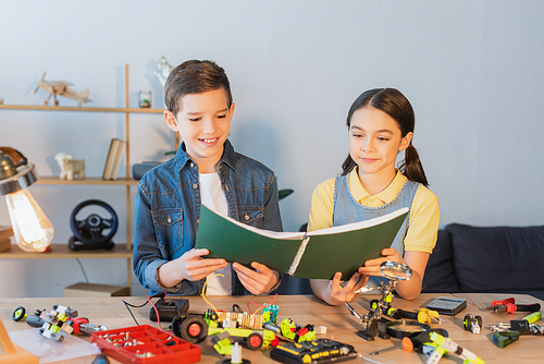 Smiling kids holding notebook near robotic model and tools at home