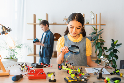 Smiling preteen child holding magnifying glass near robotics model at home