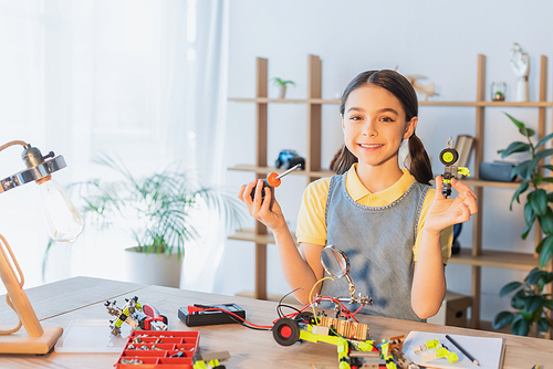 Smiling preteen girl looking at camera while holding screwdriver and robotic model at home