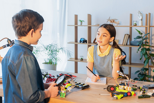 preteen girl with notebook and pencil talking to friend holding magnifier near parts of robotics model