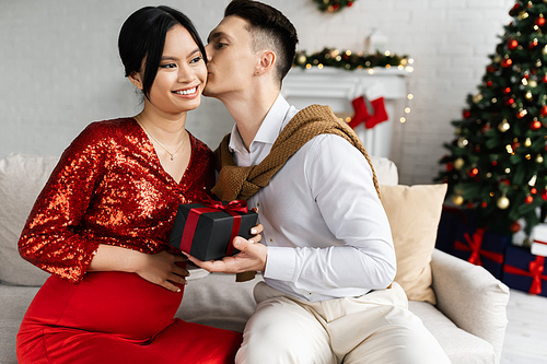 pregnant asian woman in elegant clothes smiling near husband with Christmas present kissing her on couch at home