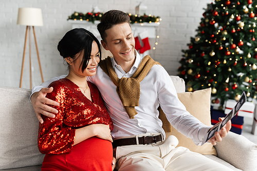happy man with ultrasound scan hugging pregnant asian woman in elegant clothes near blurred Christmas tree