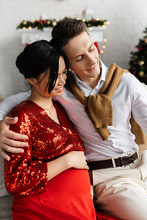 pleased man embracing pregnant asian wife in red and shiny clothes while celebrating Christmas at home