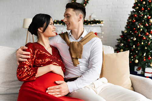 pregnant asian woman and husband in festive clothes looking at each other on couch near blurred Christmas tree