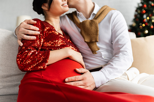 cropped view of man embracing pregnant wife in red and shiny clothes while celebrating Christmas at home