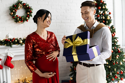 pregnant asian woman smiling near happy husband opening gift box in living room with Christmas decor