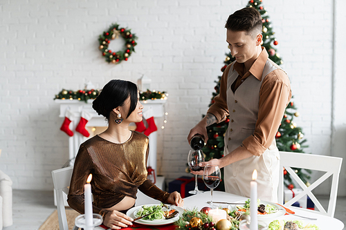 elegant man pouring wine near asian wife during romantic supper on Christmas celebration