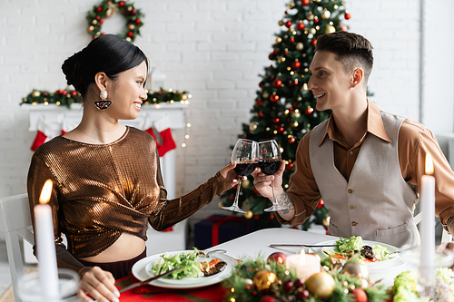 cheerful interracial couple clinking wine glasses during romantic supper in living room with Christmas decor