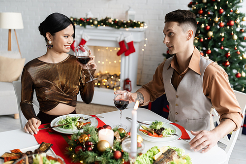 happy asian woman holding wine glass during romantic supper with husband near blurred Christmas tree