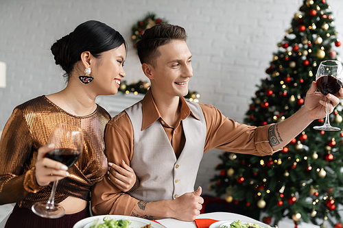 smiling man toasting with wine glass while celebrating Christmas with happy asian wife