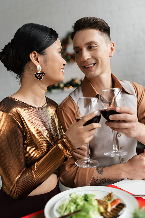 joyful multiethnic couple in elegant outfit looking at each other and clinking wine glasses during romantic supper