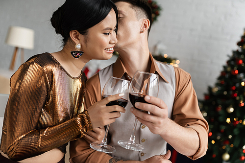 young man kissing happy and elegant asian wife and clinking wine glasses while celebrating Christmas