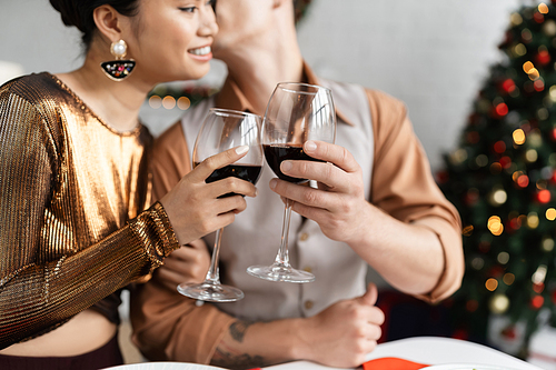 smiling asian woman in shiny blouse clinking wine glasses with blurred husband during Christmas celebration