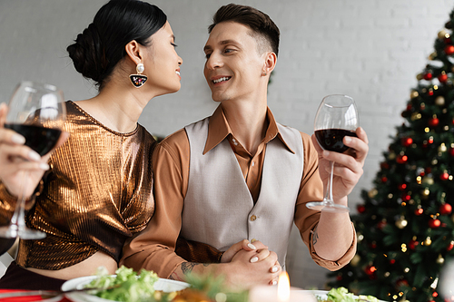 interracial couple in festive clothes holding hands and smiling at each other while holding wine glasses