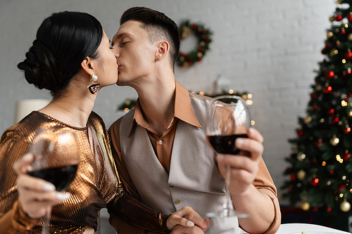 young and elegant multiethnic couple with wine glasses holding hands and kissing during Christmas celebration