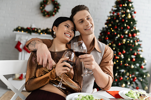 cheerful and elegant multiethnic couple clinking wine glasses during Christmas supper at home