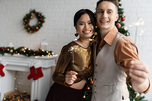 cheerful and elegant interracial couple holding shiny sparklers and looking at camera during Christmas celebration