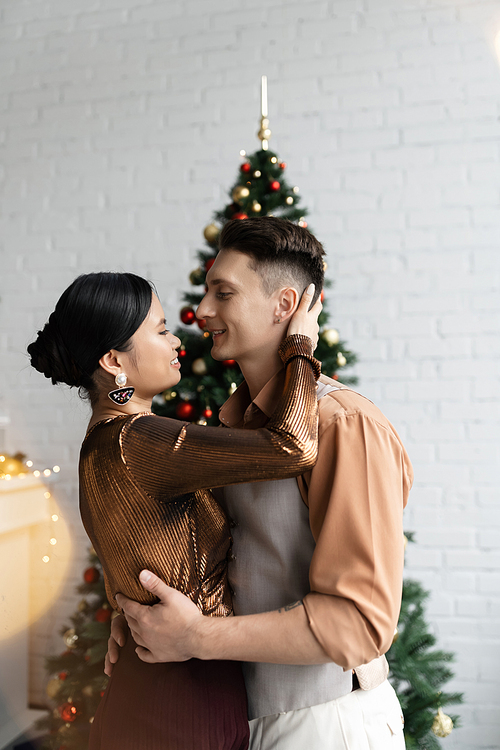 happy man and smiling asian woman in festive outfits hugging near Christmas tree