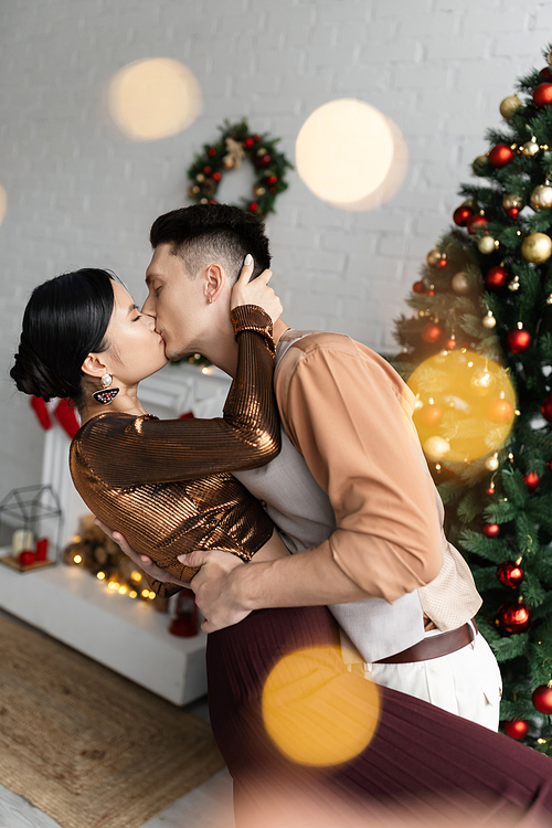 interracial couple in festive outfits kissing while standing near Christmas tree