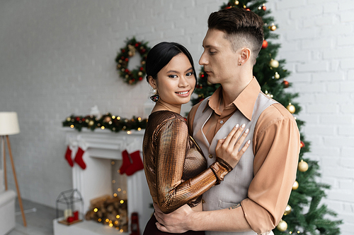 man hugging cheerful asian wife in festive outfit while standing near Christmas tree