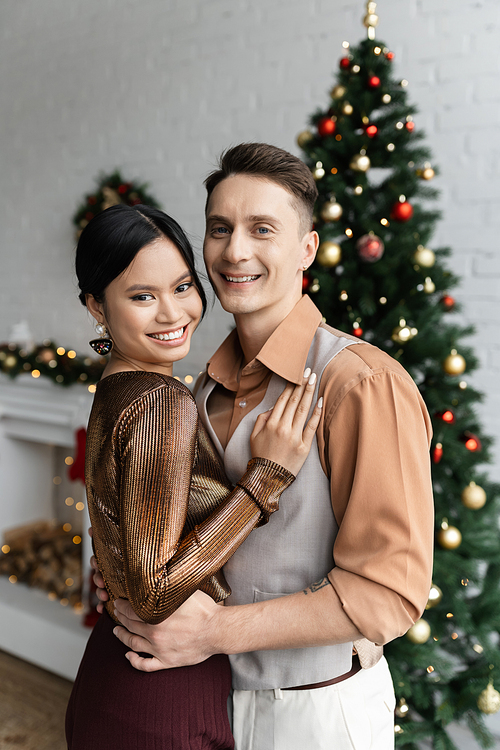 cheerful asian woman and man in festive outfits hugging near Christmas tree