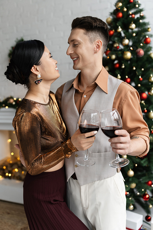 cheerful multiethnic couple looking at each other while clinking glasses of red wine near Christmas tree