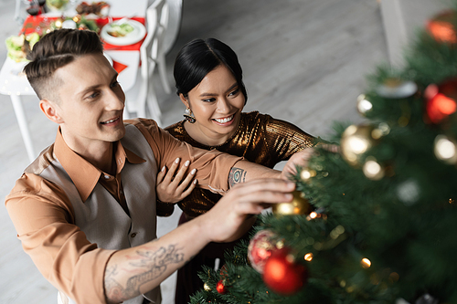 high angle view of smiling interracial couple decorating Christmas tree with shiny baubles